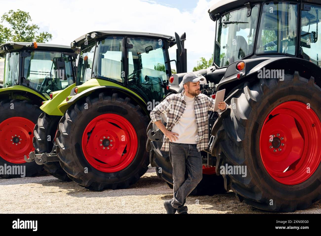 Händler oder Lieferant von Landmaschinen. Kaukasischer Mann im karierten Hemd steht neben Traktoren und benutzt ein digitales Tablet. Stockfoto