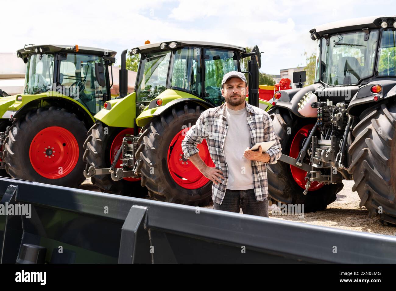 Kaukasischer Erwachsener mit digitalem Tablet in der Hand steht neben einem Bau- oder landwirtschaftlichen Traktor bei einem Maschinenhändler. AG-Gerätehändler oder -Zulieferer Stockfoto