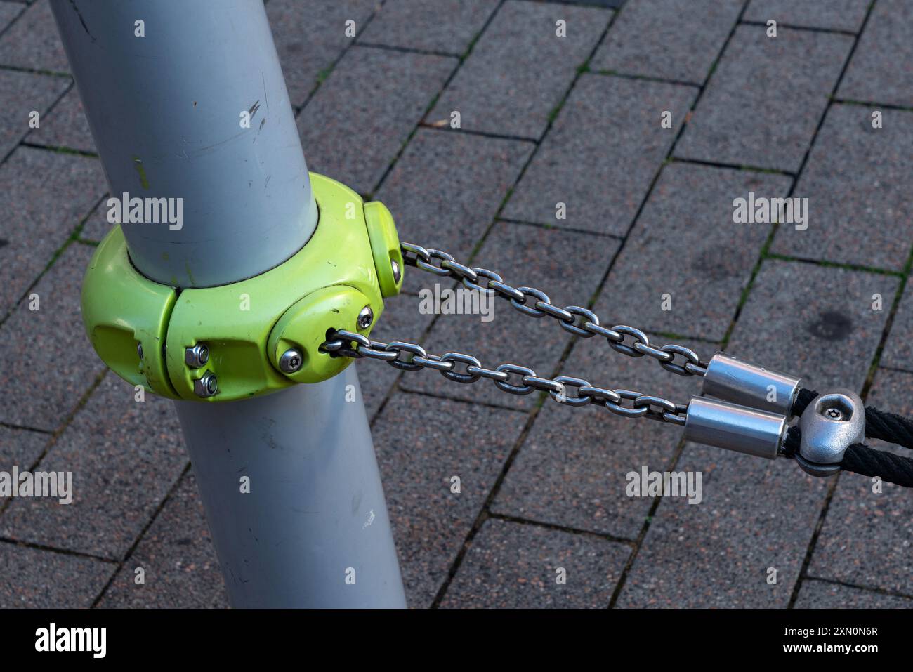 Ein hellgrüner Verbindungsstück sichert eine Stahlkette an einem Stab auf einem gepflasterten Bürgersteig und zeigt ein funktionales Design in urbanen Umgebungen. Stockfoto