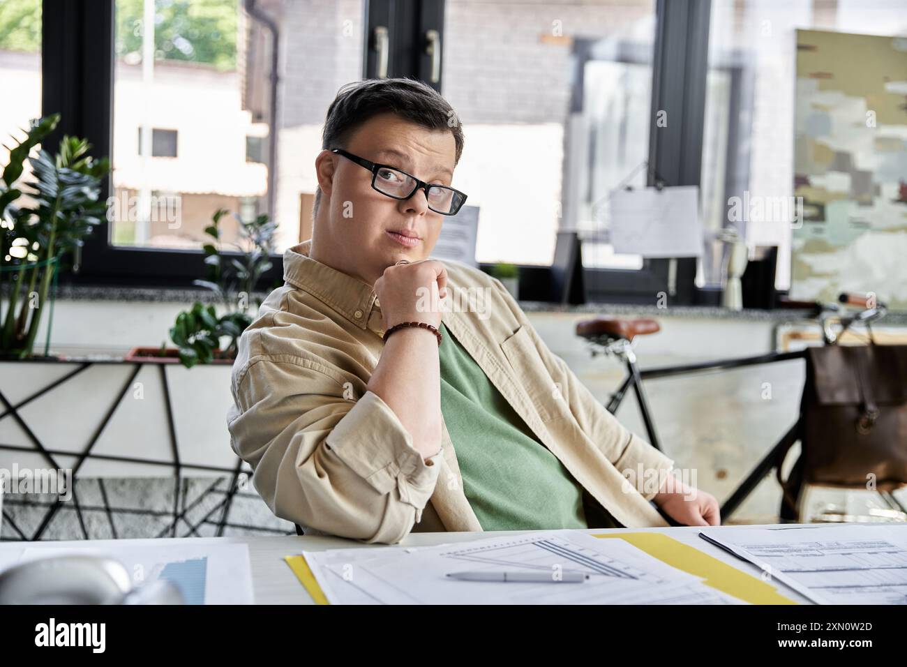 Ein junger Mann mit Down-Syndrom sitzt an einem Schreibtisch und legt nachdenklich sein Kinn auf seine Hand. Stockfoto