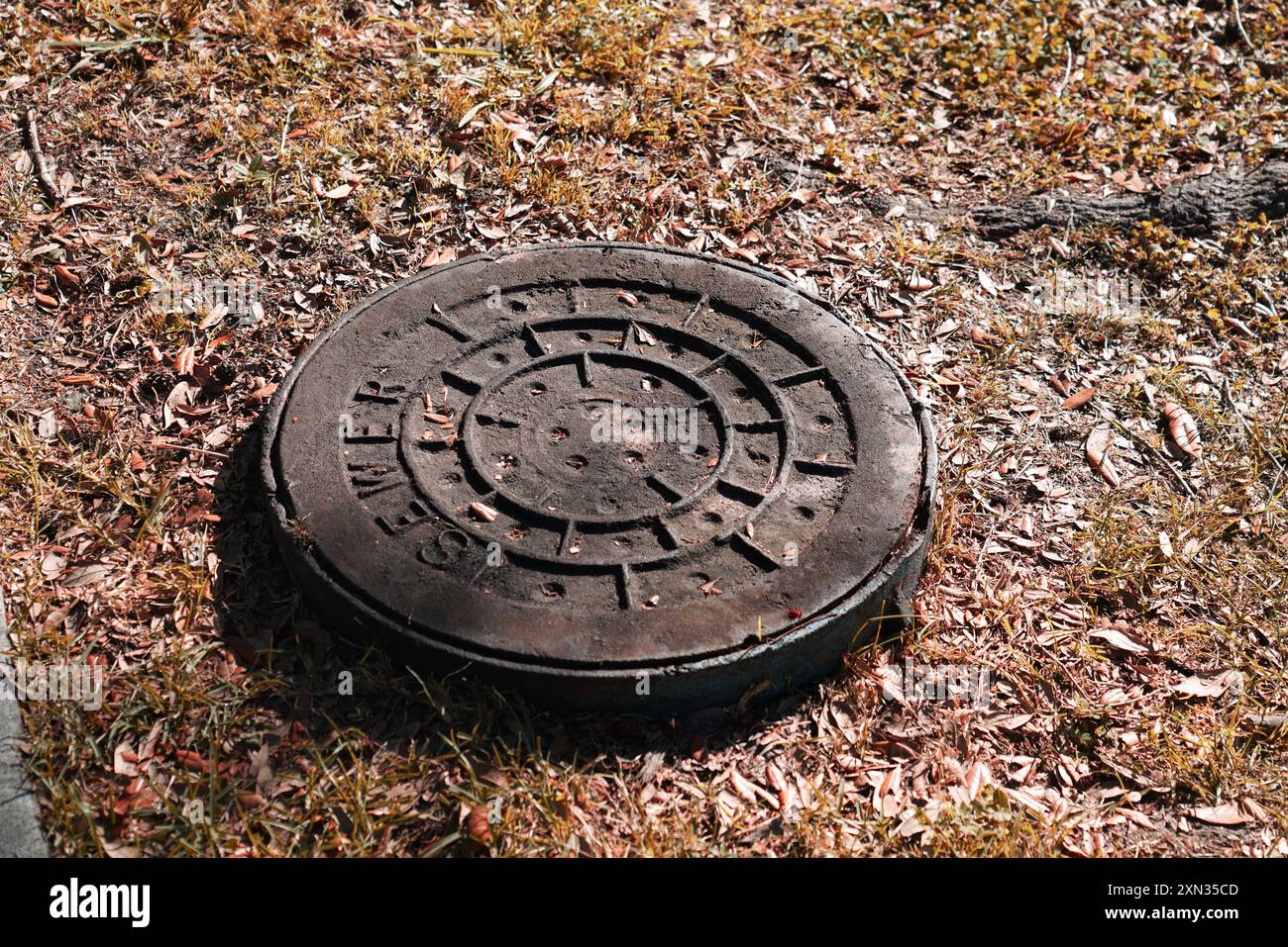 Eine Mannlochabdeckung auf trockenem, grasbewachsenem Boden, die städtische Infrastruktur und Details zeigt. Stockfoto