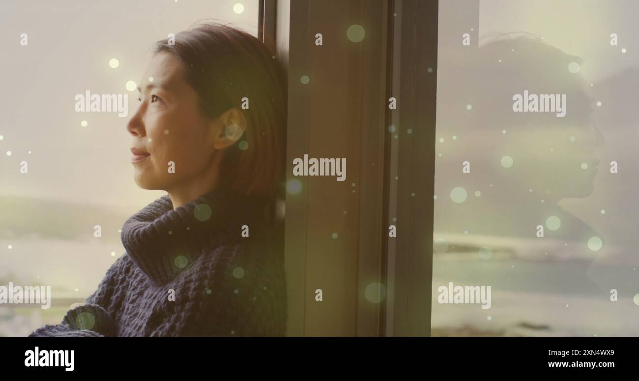 Bild von Lichtflecken über asiatischer Frau, die zu Hause durch das Fenster schaut Stockfoto