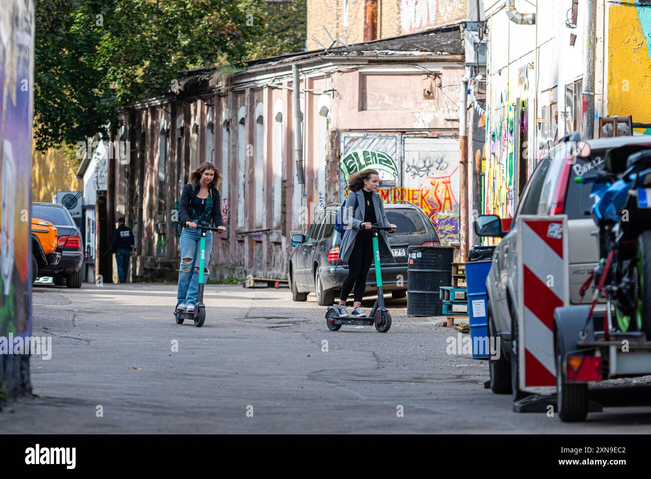 Riga, Lettland - 30. September 2020: Zwei Mädchen fahren in einer lebhaften urbanen Gasse, umgeben von Graffiti, Motorroller. Stockfoto