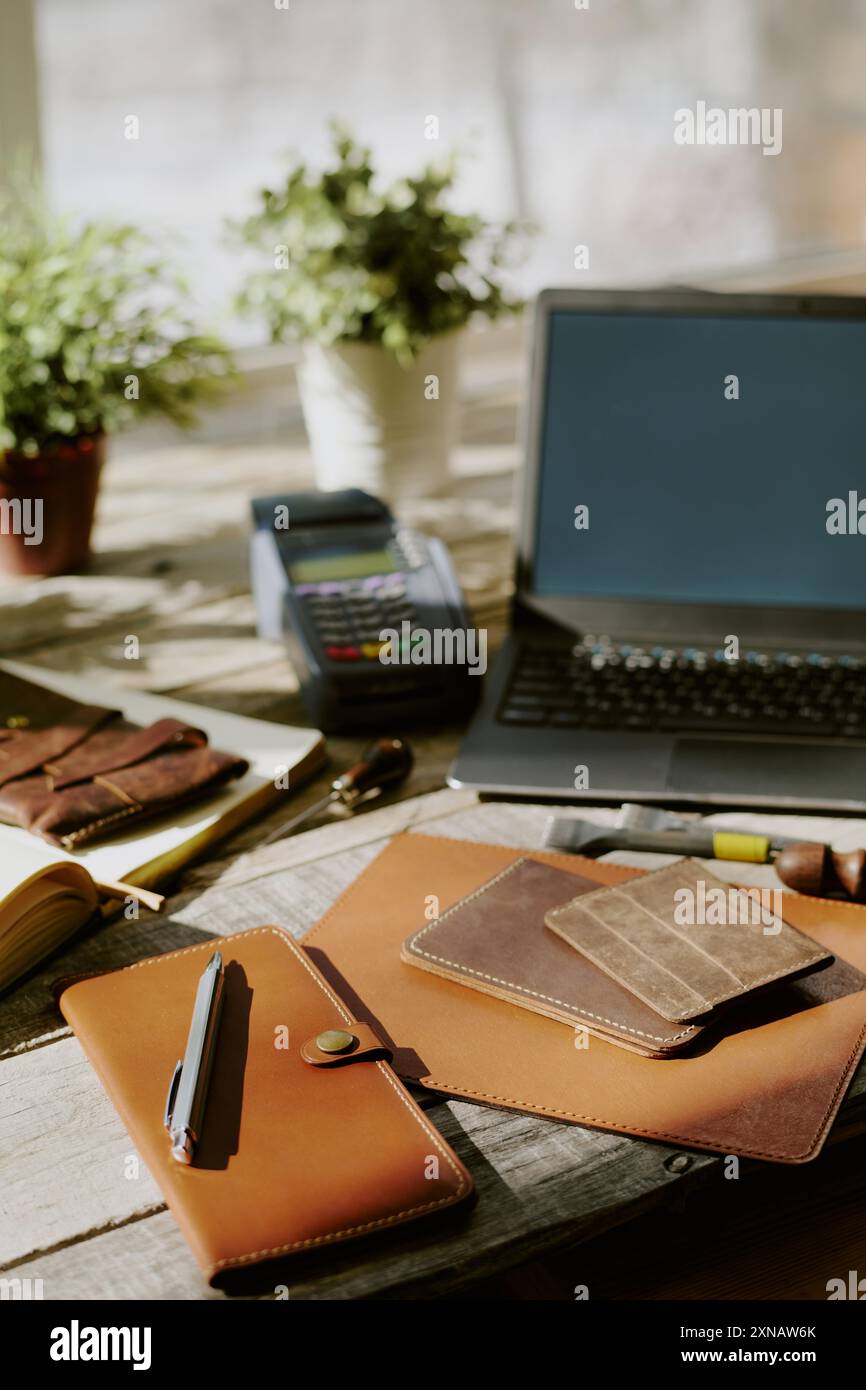 Handgefertigte Ledernotizbücher und Kartenhalter auf altem Holztisch, Laptop und Zahlungsterminal im Hintergrund Stockfoto