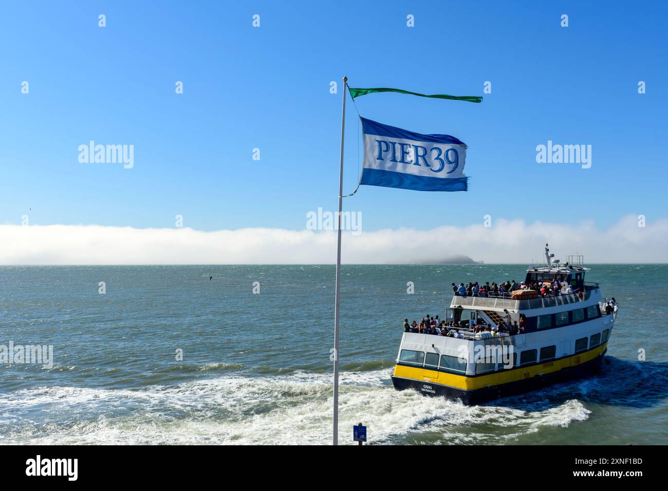 Die Flagge von Fisherman's Wharf am Pier 39 flattert über einer Fähre Osky von Blue and Gold Fleet Company. Nebelige Insel Alcatraz im Hintergrund - San Fra Stockfoto