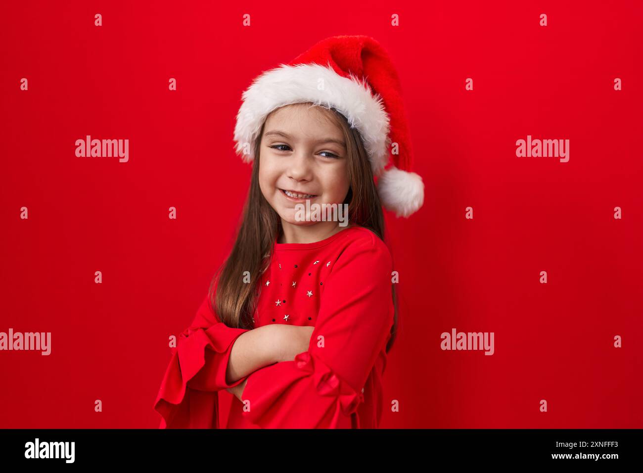 Entzückendes hispanisches Mädchen, Arme gekreuzt, trägt einen weihnachtsmütze über rotem Hintergrund. Ihr fröhliches, strahlendes Lächeln strahlt auf die Kamera. Was für ein Bündel positiver Ergebnisse Stockfoto