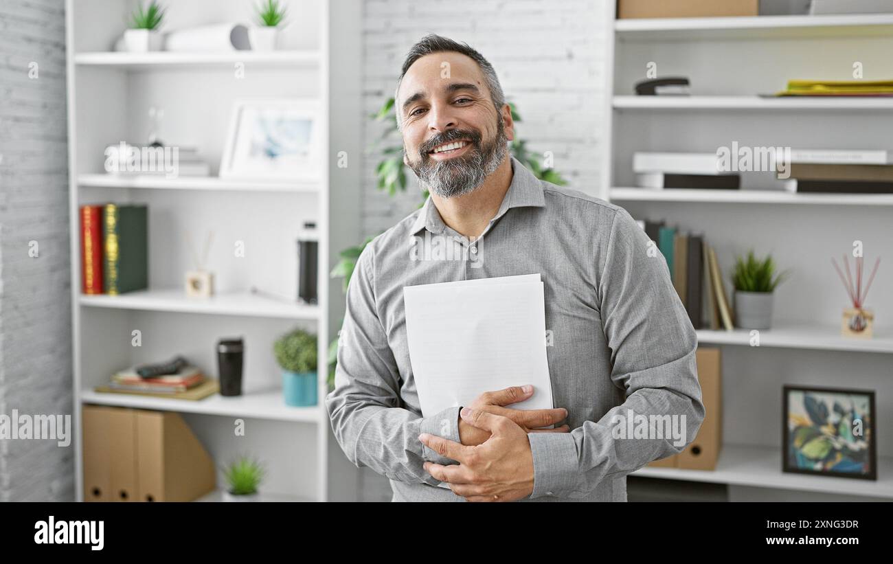 Gutaussehender ranghanischer Mann mit grauem Bart, der im modernen Büro lächelt und Dokumente hält Stockfoto