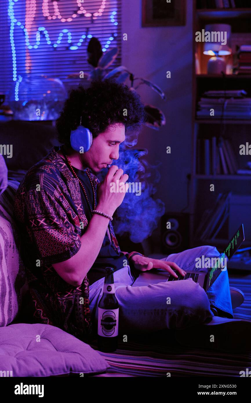 Entwickler mit lockigen Haaren, der süßen Vape inhaliert und Codes auf dem Laptop überprüft Stockfoto