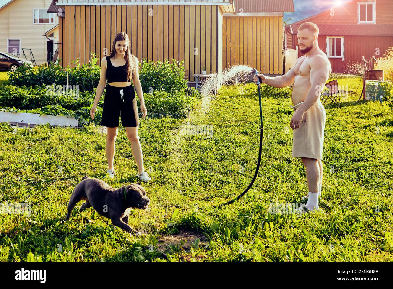 Weißer Mann in Shorts und ohne Hemd spielt mit dem Hund, leitet den Wasserstrahl vom Schlauch zum Tier, junge Frau sieht Spaß zu. Stockfoto