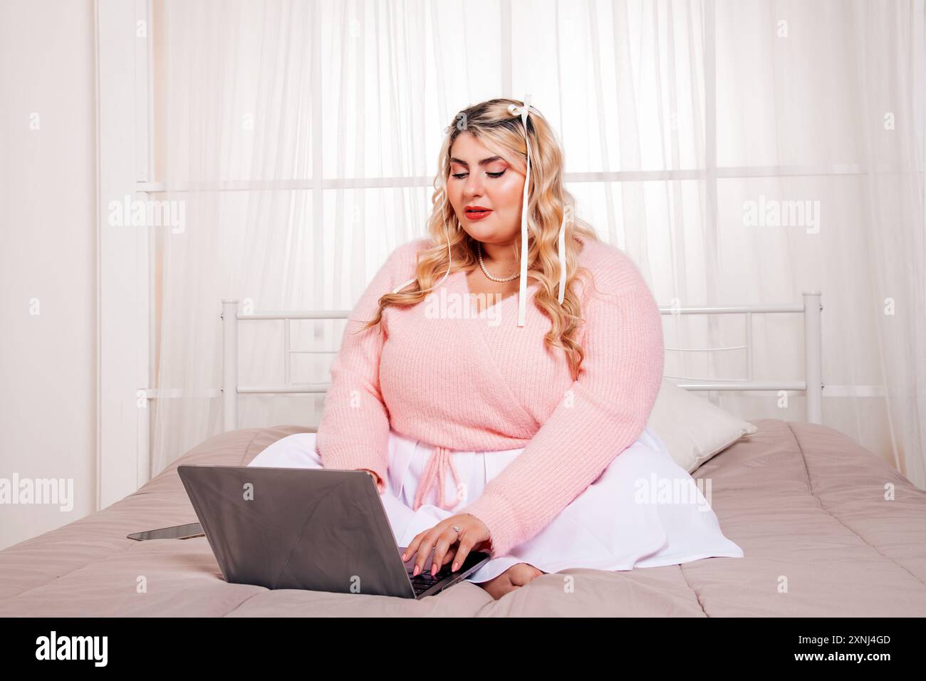 Frau in Übergröße, die von zu Hause aus arbeitet, bequem auf ihrem Bett sitzt und auf ihrem Laptop schreibt Stockfoto