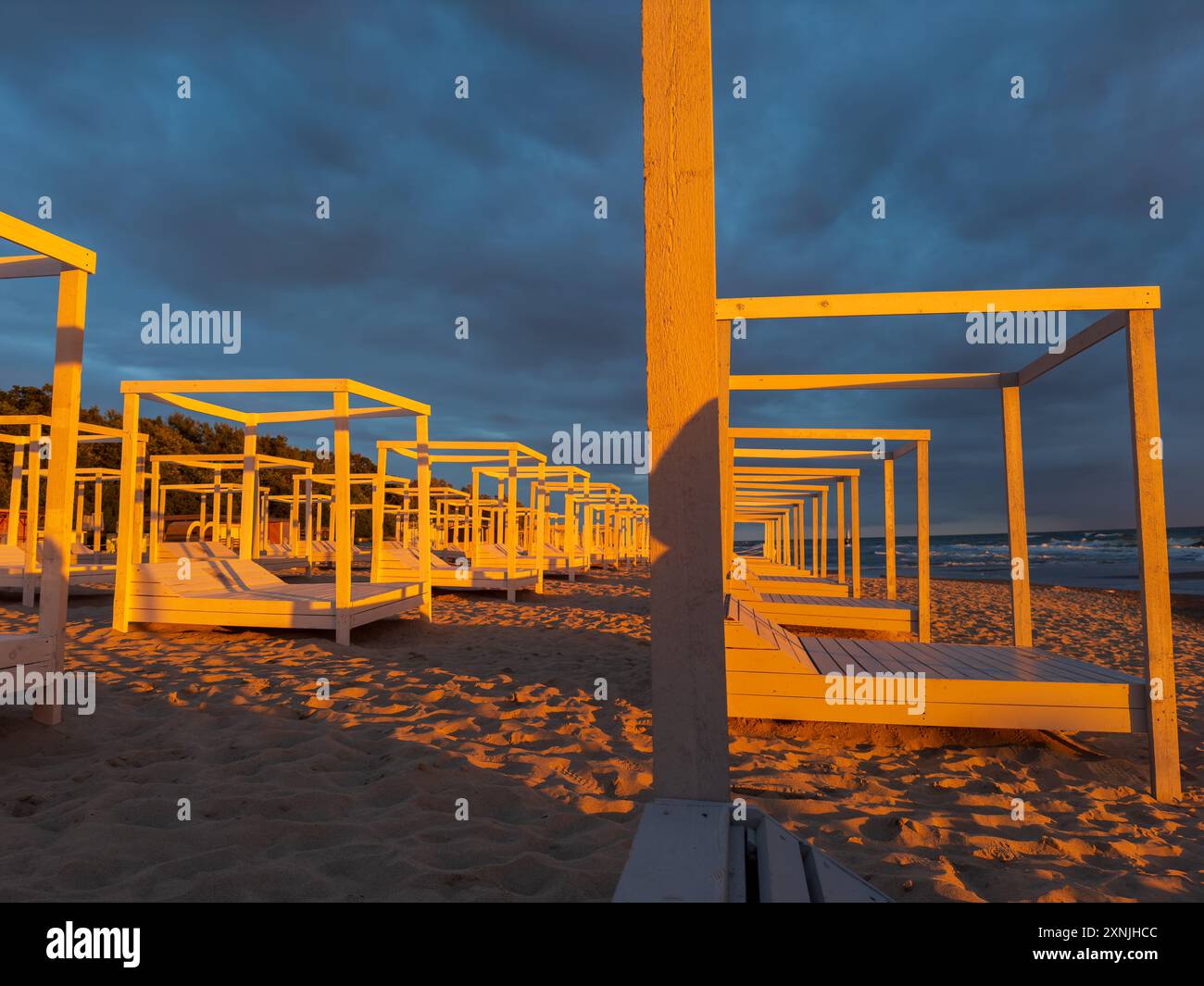 Eine Reihe leerer hölzerner Strandliegen am Sandstrand mit einem dramatischen bewölkten Himmel während des Sonnenuntergangs, die eine ruhige und friedliche Atmosphäre schaffen. Stockfoto