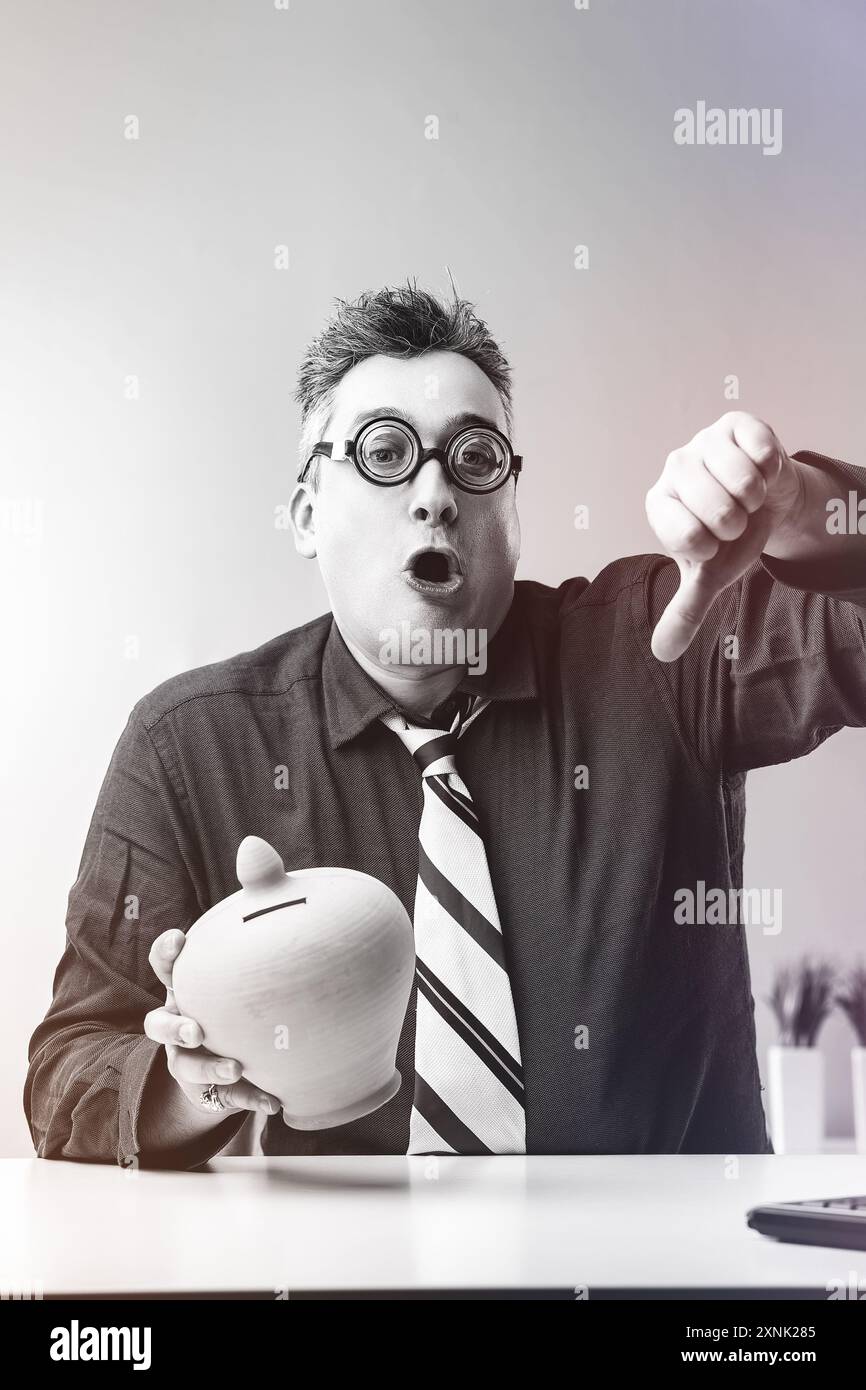 Ein Geschäftsmann hält eine Sparkasse und drückt mit schockiertem Ausdruck die Daumen nach unten, was auf finanzielle Probleme hindeutet Stockfoto