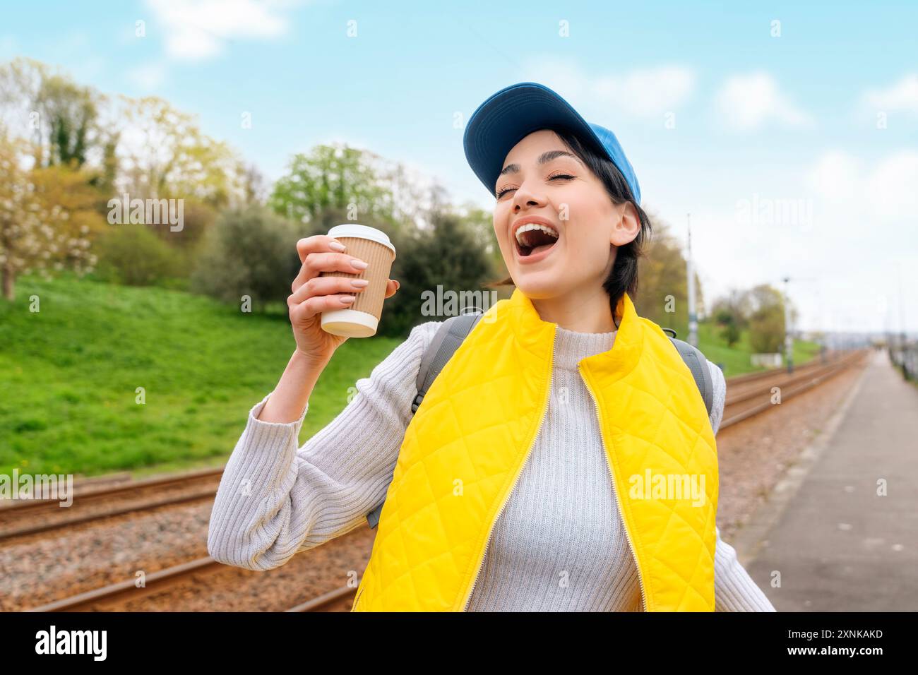 Glückliche junge Frau in gelber Weste und blauer Mütze, die Kaffee zum Mitnehmen, Tee auf der Straße trinkt und Spaß hat. Lifestyle-Konzept Stockfoto