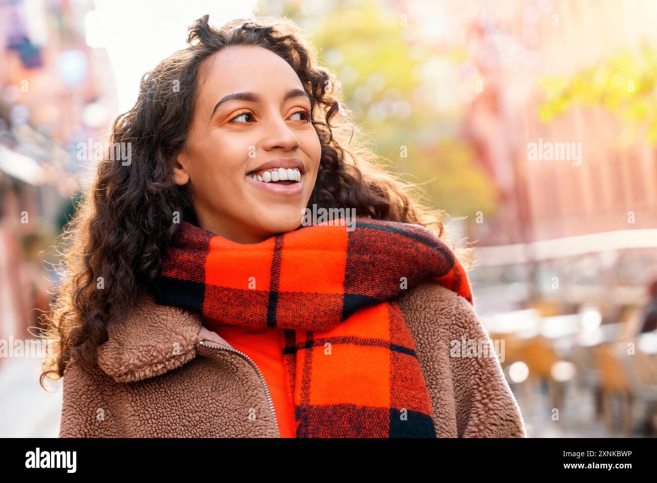 Eine fröhliche Frau, die eine Tasse Kaffee hält. Eine lächelnde, lockige Brünette in einem Pullover, die auf eine Straßenbahn wartet. Stockfoto