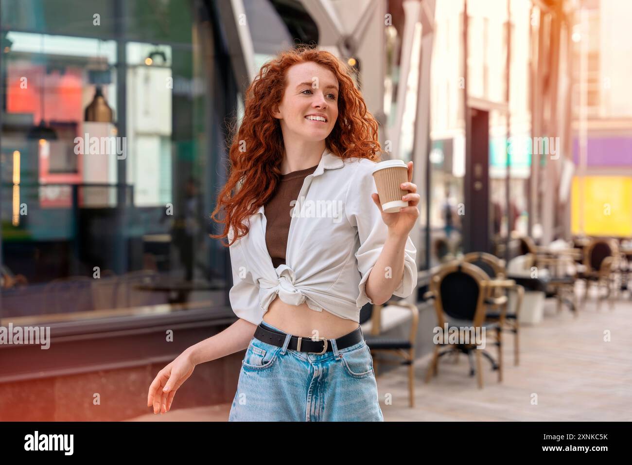 Glückliche junge rothaarige Frau in Jeans und weißem Hemd, die auf der Straße Kaffee zum Mitnehmen trinkt und Spaß hat. Lifestyle-Konzept Stockfoto