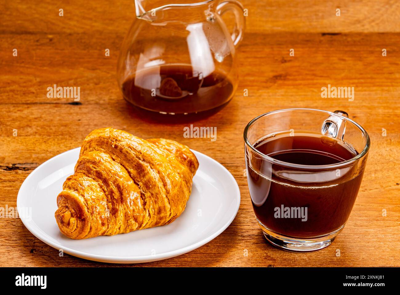 Blick auf frisch gebackenes köstliches französisches Croissant in weißer Keramikschale mit einer Tasse schwarzen Kaffee und einer transparenten Glaskaffeekanne auf Woo Stockfoto