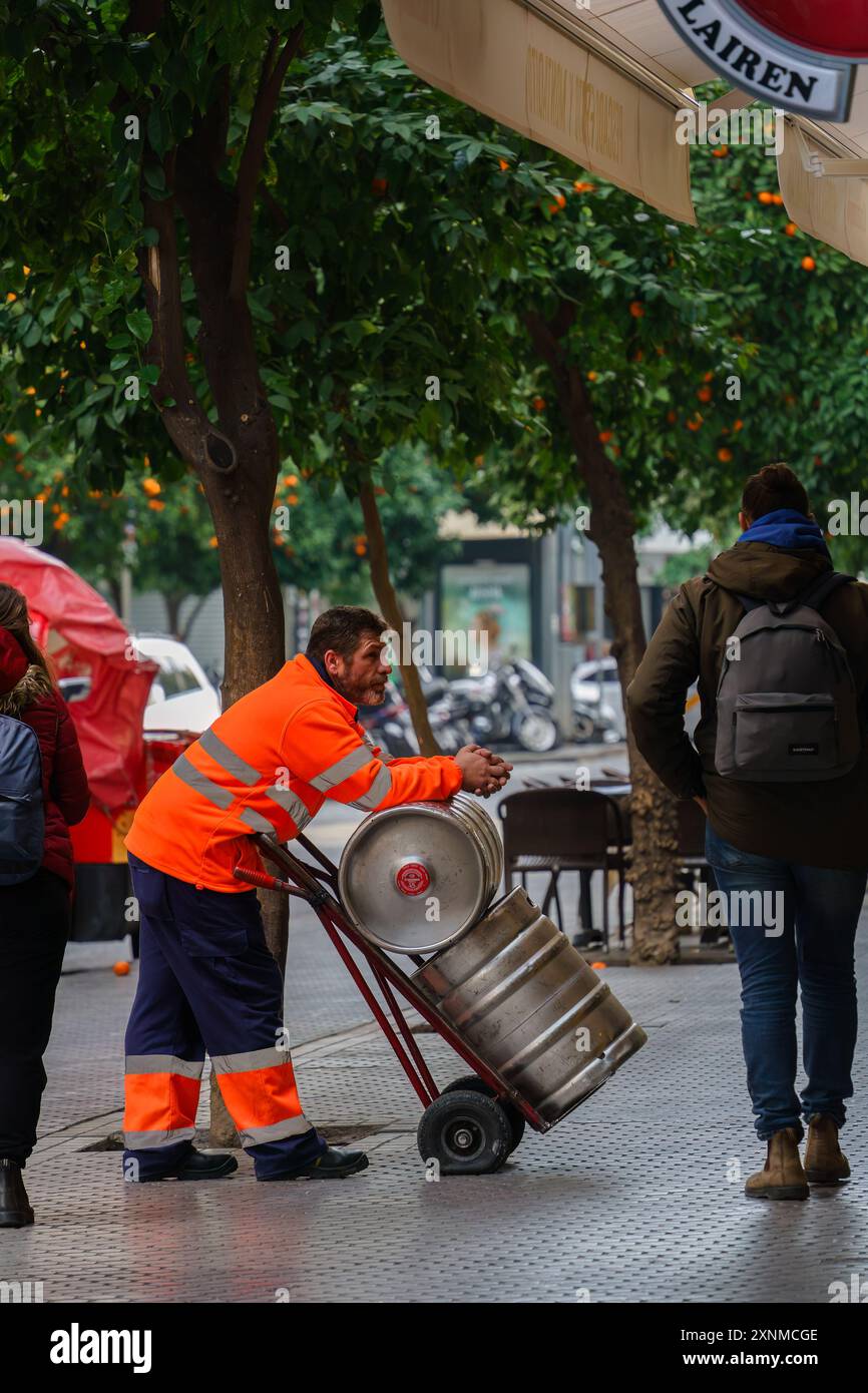 Sevilla, Spanien. 5. Februar 2024: Mann in orangefarbener Arbeitskleidung, der sich mit zwei Fässern auf einen Handwagen stützt Stockfoto