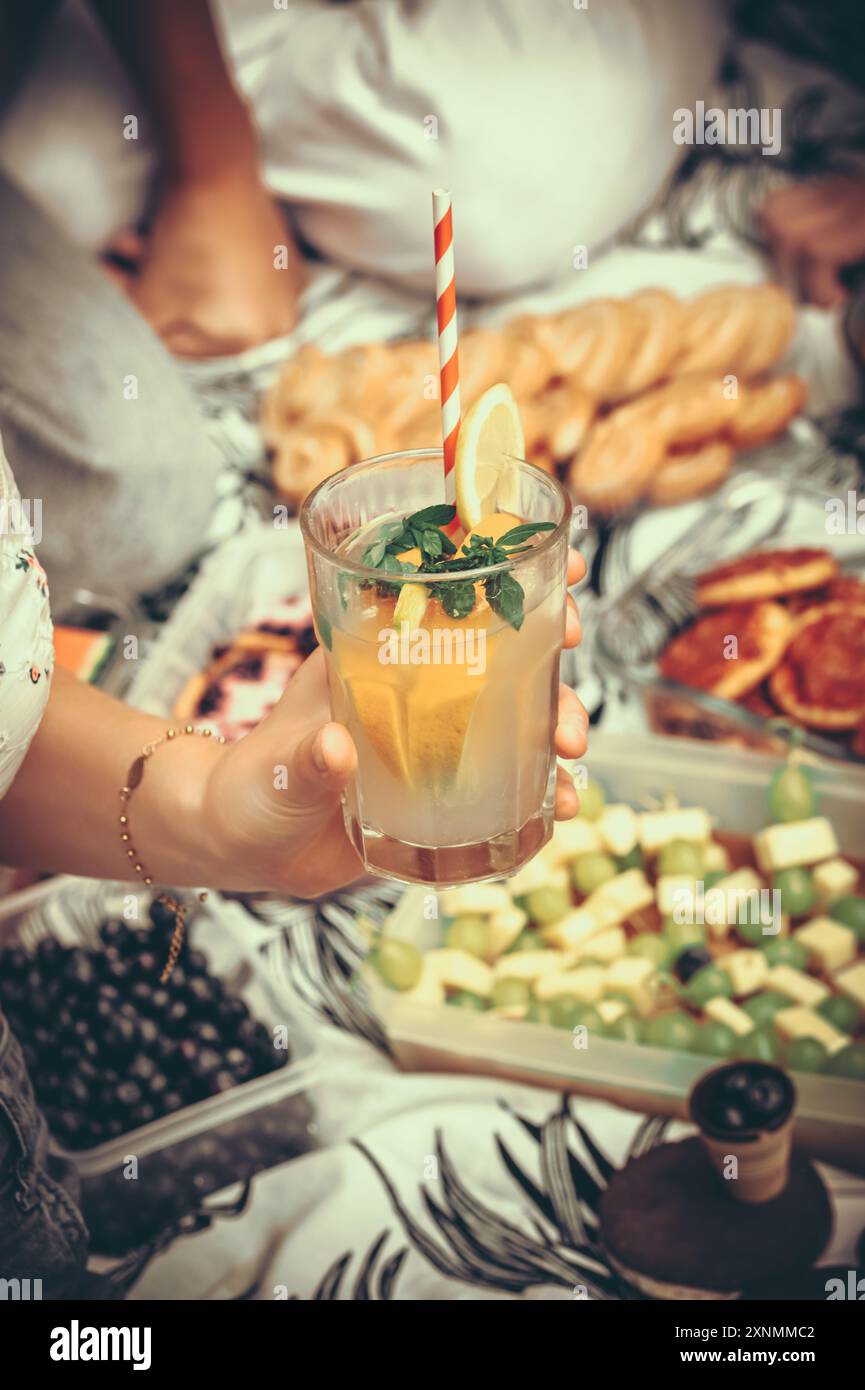Eine weibliche Hand hält einen Cocktail mit Zitronen bei einem Picknick im Sommer Stockfoto