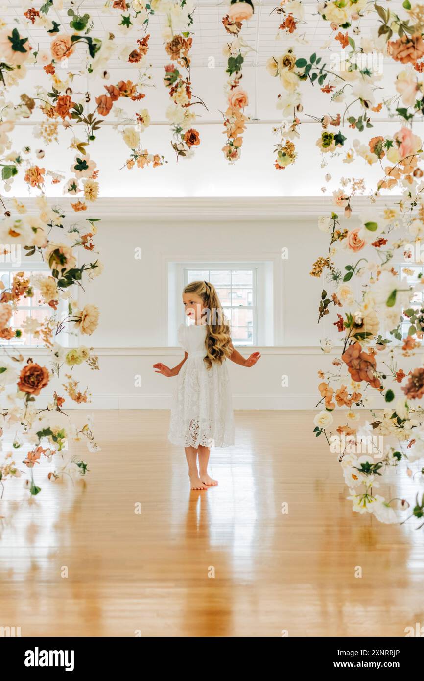 Blondes Mädchen in weißem Kleid steht ernsthaft zwischen hängenden Blumen Stockfoto