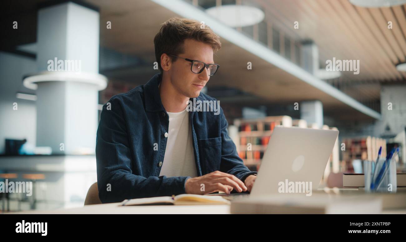 Konzentrierter junger kaukasischer männlicher Student, der an seinem Laptop in einer modernen Bibliothek arbeitet. Mann beschäftigte sich mit akademischer Forschung mit Books Around, fokussiert auf Bildschirm. Stockfoto