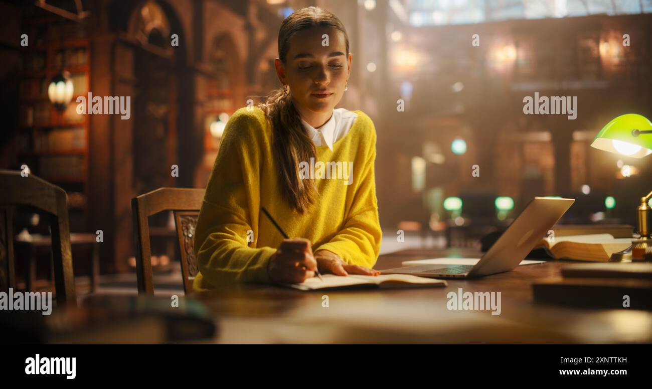 Fokussierte junge Frau, die in einer Vintage-Bibliothek, umgeben von Büchern, mit einem Laptop für Forschung und Schreiben in Notebook studiert. Ruhiges akademisches Setting unterstreicht das Engagement für Lernen und Bildung Stockfoto