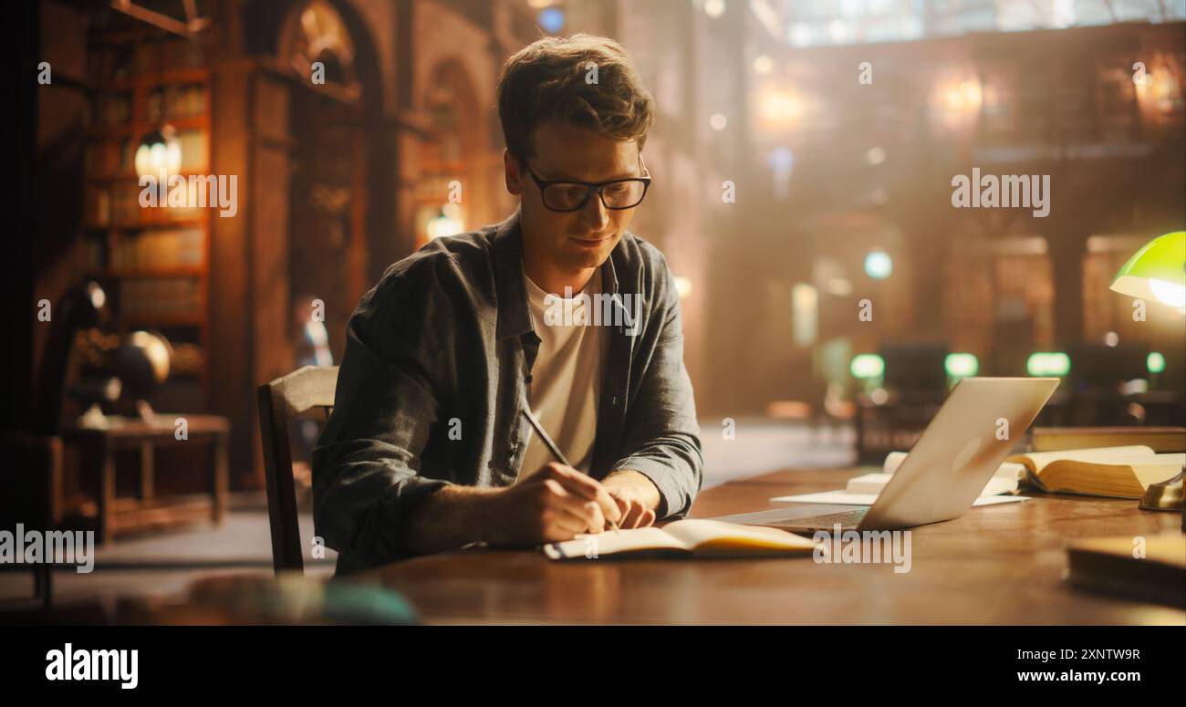 Konzentrierter junger Mann, der in Notizbuch an einem Vintage Library Desk schreibt, mit Laptop für Online-Forschung geöffnet. Die warme, atmosphärische Beleuchtung unterstreicht die klassische Architektur, die ihn umgibt Stockfoto