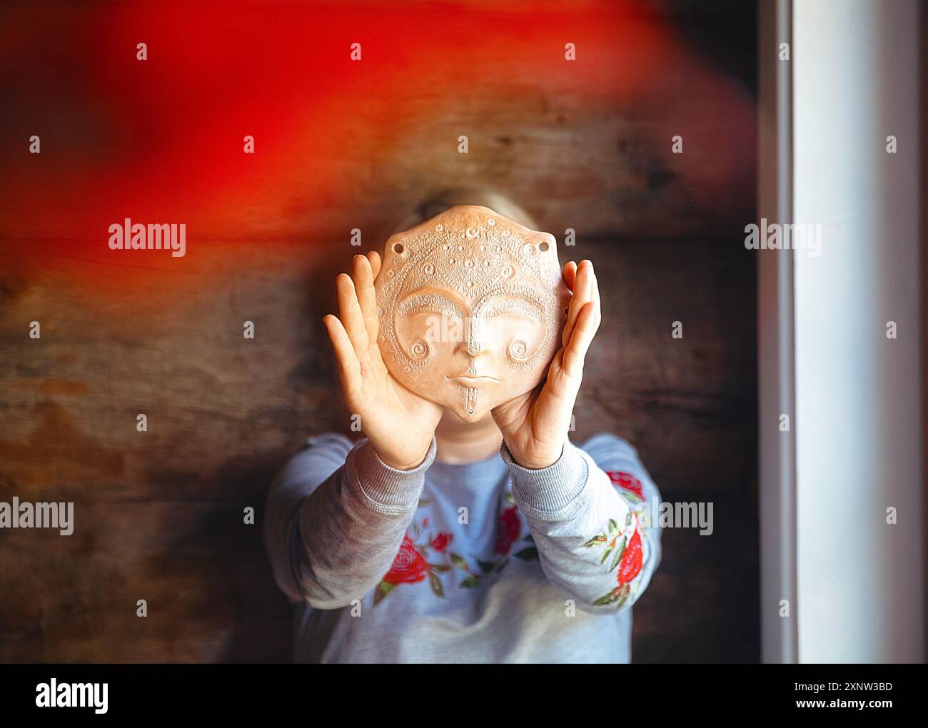 Eine Frau hält eine Tonmaske, die sein Gesicht bedeckt, mit komplizierten, skurrilen Mustern. Der Hintergrund hat eine rustikale, hölzerne Textur Stockfoto