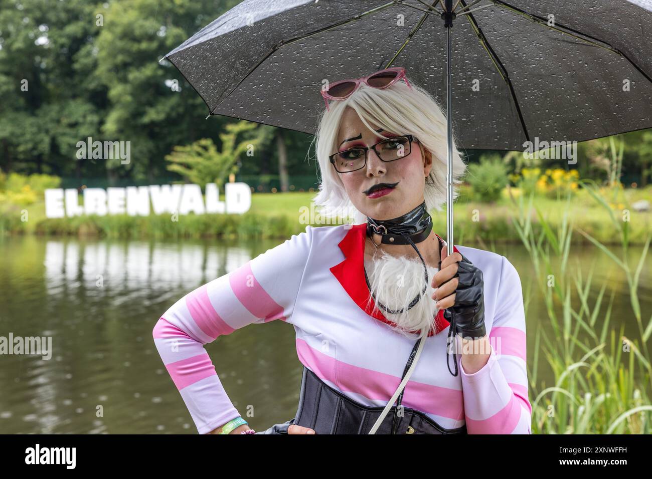 02. August 2024, Brandenburg, Cottbus: Tessa Wilken aus Haselünne im Emsland steht beim Elbenwald Festival am Teich im Spreeauenpark im Regen. Sie verkleidet sich als Angel Dust aus der Zeichentrickserie „Hazbin Hotel“ und nimmt zum ersten Mal am Elbenwald Festival Teil. Das Elbenwald Festival ist in erster Linie ein Treffpunkt für Fans populärer Science-Fiction- und Fantasy-Serien wie Harry Potter, Star Wars, Lord of the Rings und Marvel Comics. Foto: Frank Hammerschmidt/dpa - ACHTUNG: Nur zur redaktionellen Verwendung im Zusammenhang mit der aktuellen Berichterstattung und nur mit vollständigem Verweis t Stockfoto