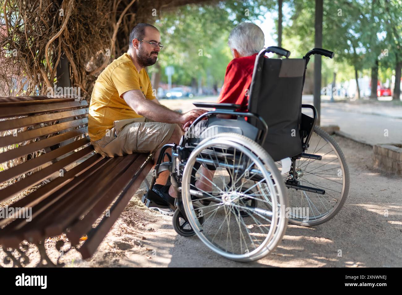 Junger Mann, der auf einer Bank sitzt und glücklich mit seiner weißhaarigen Großmutter im Rollstuhl redet. Stockfoto
