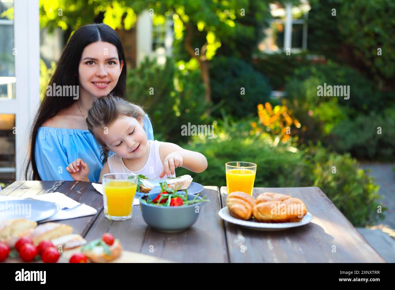 Glückliche junge Frau in Casual Wear, die am Tisch sitzt und die kleine Tochter hält, während sie gesundes Abendessen mit frischem Salat und Brot mit Orange hat Stockfoto