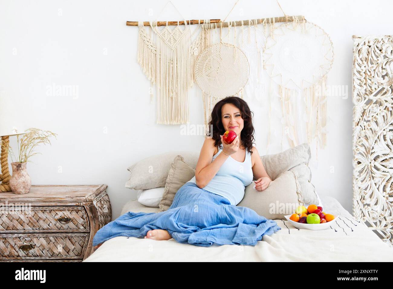 Glückliche junge Frau in der Erwartung, die roten Apfel hält und in die Kamera blickt, während sie auf dem Bett mit einem Teller Obst in einem hellen Boho-Stil mit sitzt Stockfoto