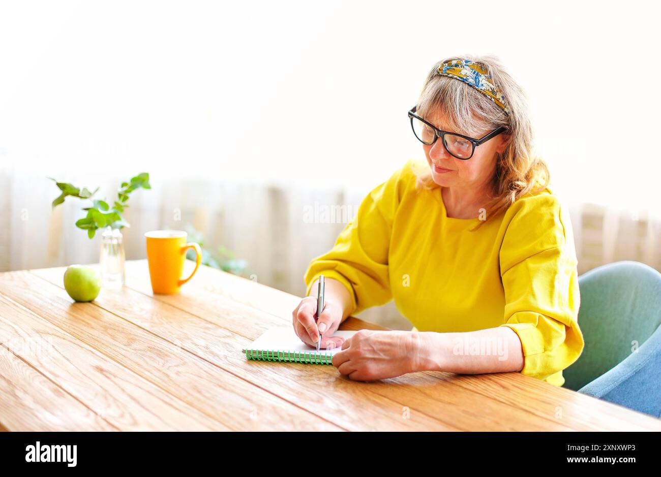 Ruhige ältere Frau in hellen Kleidern und Brillen, die zu Hause am Holztisch sitzen und im Notizbuch schreiben Stockfoto