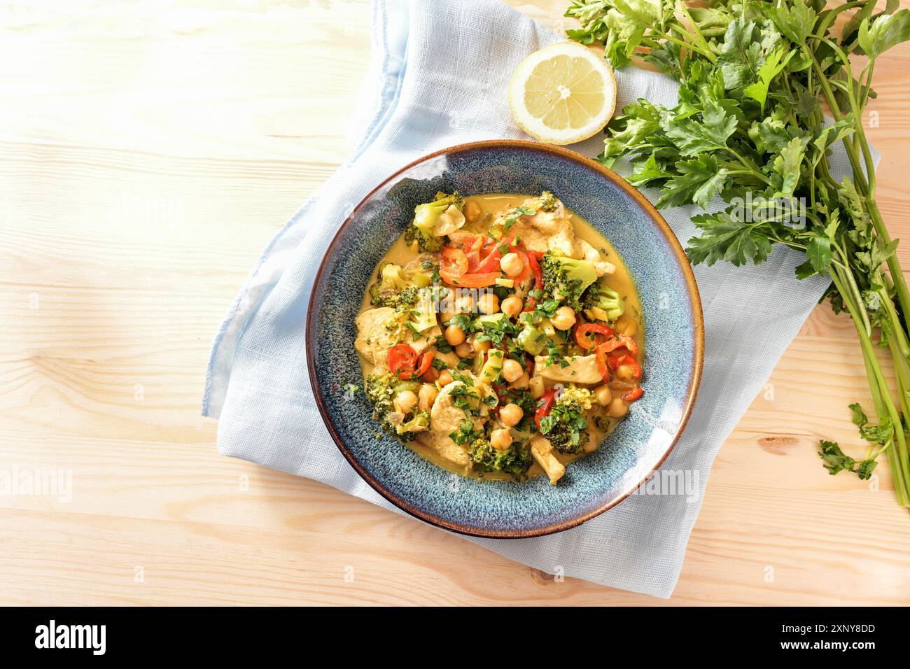 Indisches Curry aus Kichererbsen, buntes Gemüse und Hühnerfleisch in einer blauen Schüssel, Zitrone, Kräuter und Serviette auf einem hellen Holztisch, Kopierraum Stockfoto