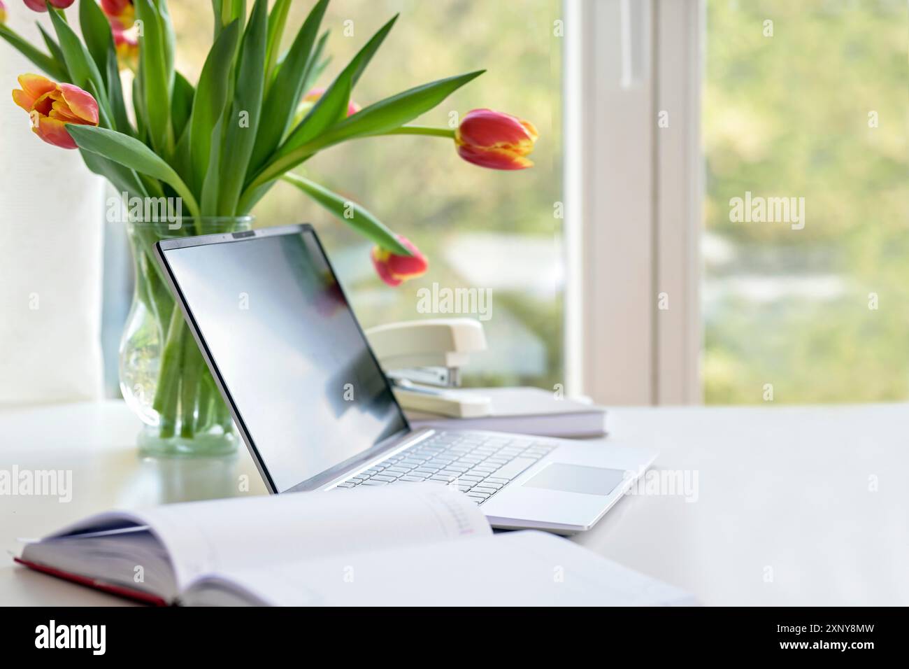 Laptop mit Organizer und Blumenstrauß aus Tulpen auf einem weißen Schreibtisch am Fenster, Business im Heimbüro, Kopierfläche, ausgewählter Fokus, schmal Stockfoto