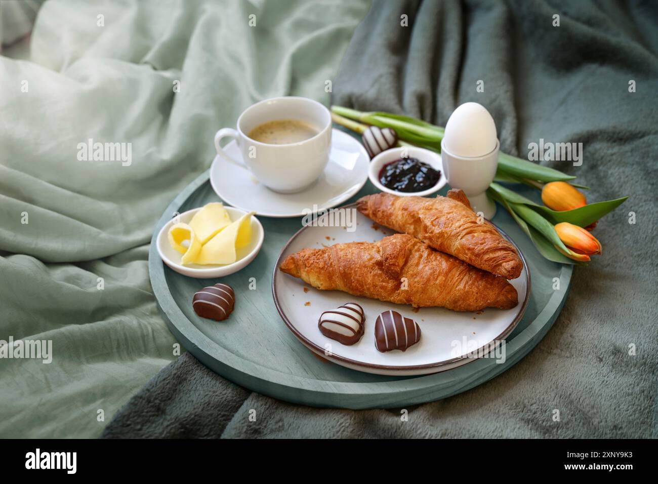 Frühstück mit Liebe im Bett, Kaffee, Croissant, Marmelade, Ei, ein paar Schokoladenherzen und Tulpen auf einem Tablett auf graugrüner Bettwäsche bei einem romantischen Stockfoto