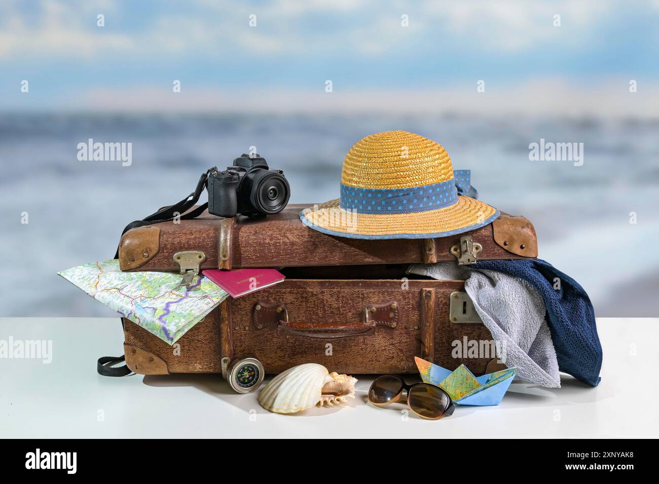 Vintage-Koffer mit Reiseaccessoires für den Strandurlaub wie Strohhut, Fotokamera, Handtücher, Reisepass, Karte und Sonnenbrille, verschwommenes Meer Stockfoto