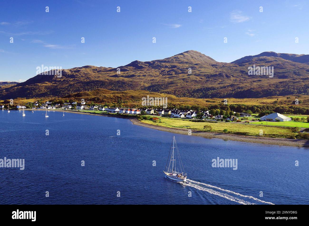 Segelboot auf dem Meer neben einem Dorf und sanften Hügeln unter blauem Himmel, Herbst, Oktober, Highlands, Kyle of Lochalsh, Schottland, Vereinigtes Königreich Stockfoto
