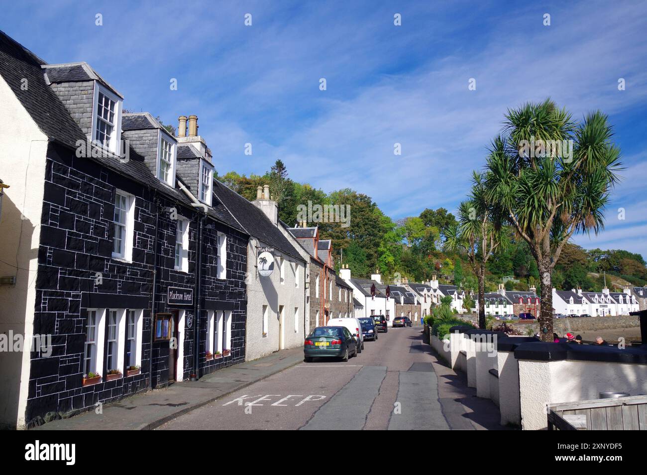 Eine ruhige Straße in einer Küstenstadt mit Häusern, Palmen und geparkten Autos unter blauem Himmel, Oktober, Plockton, Highlands Schottland, Großbritannien Stockfoto