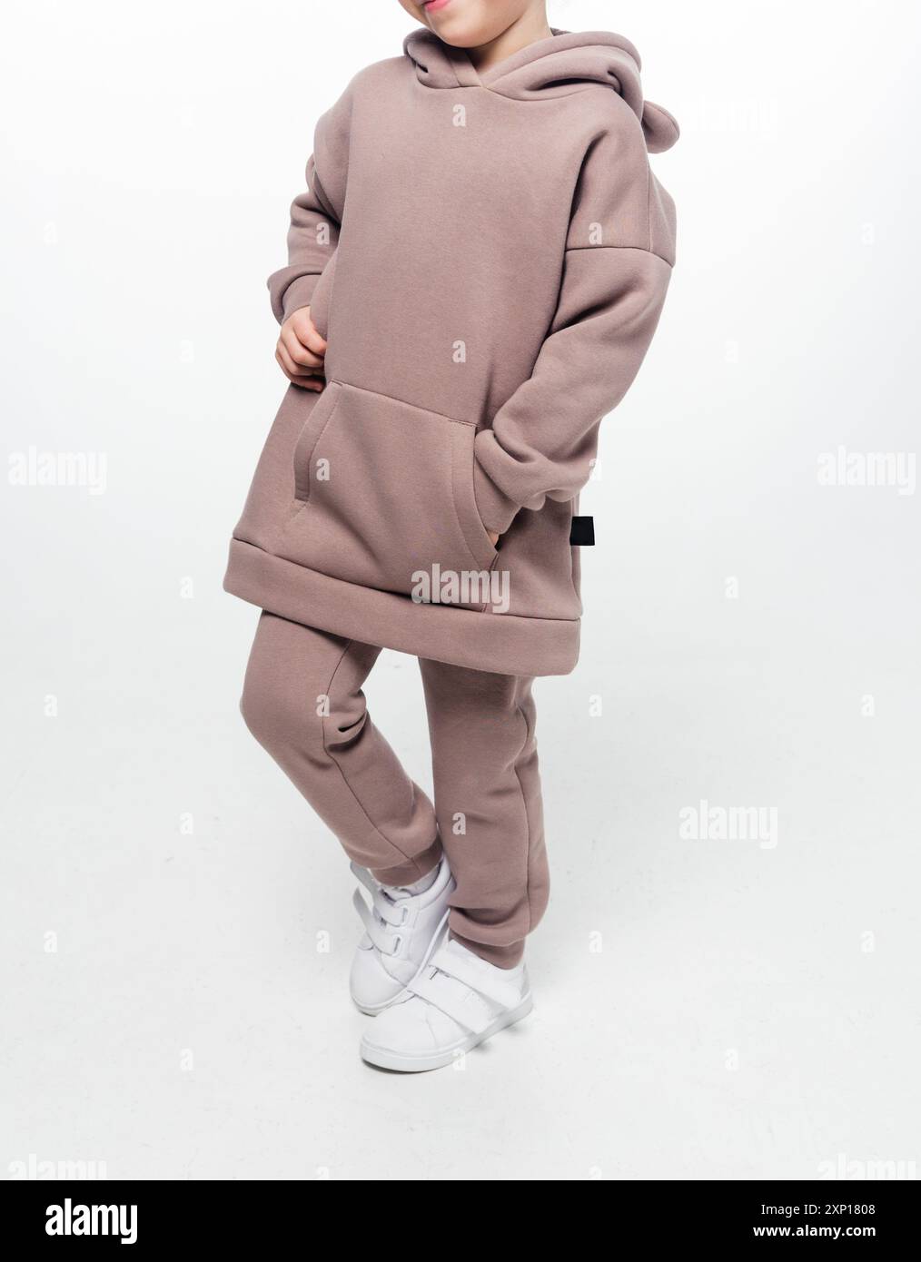 Ein junges Mädchen in braunem Hoodie und passendem Hosenset, posiert vor weißem Hintergrund. Vollständiges Outfit sichtbar. Stockfoto