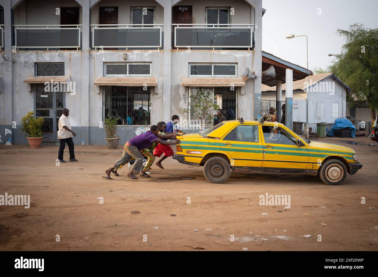 Eine Gruppe von Menschen, die gemeinsam ein kaputtes gelbes Taxi auf eine staubige Straße in Gambia schieben, um die Bemühungen und die Unterstützung der Gemeinschaft hervorzuheben. Stockfoto