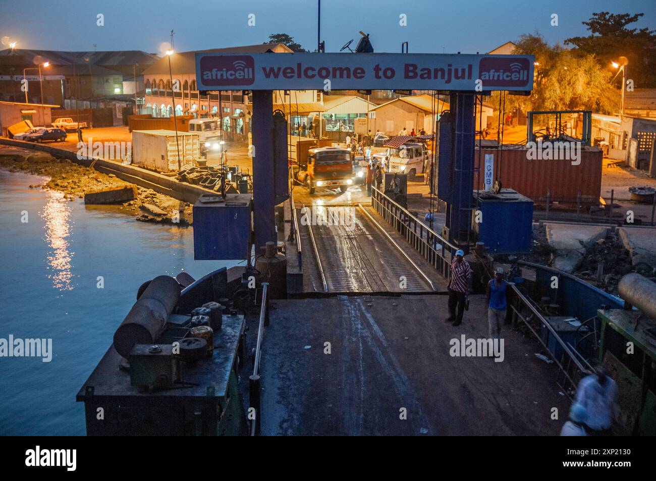 Nächtlicher Blick auf eine Fähre, die am Hafen von Banjul in Gambia ankommt, mit Fahrzeugen und Arbeitern an Bord, beleuchtet durch warme Straßenlaternen. Stockfoto