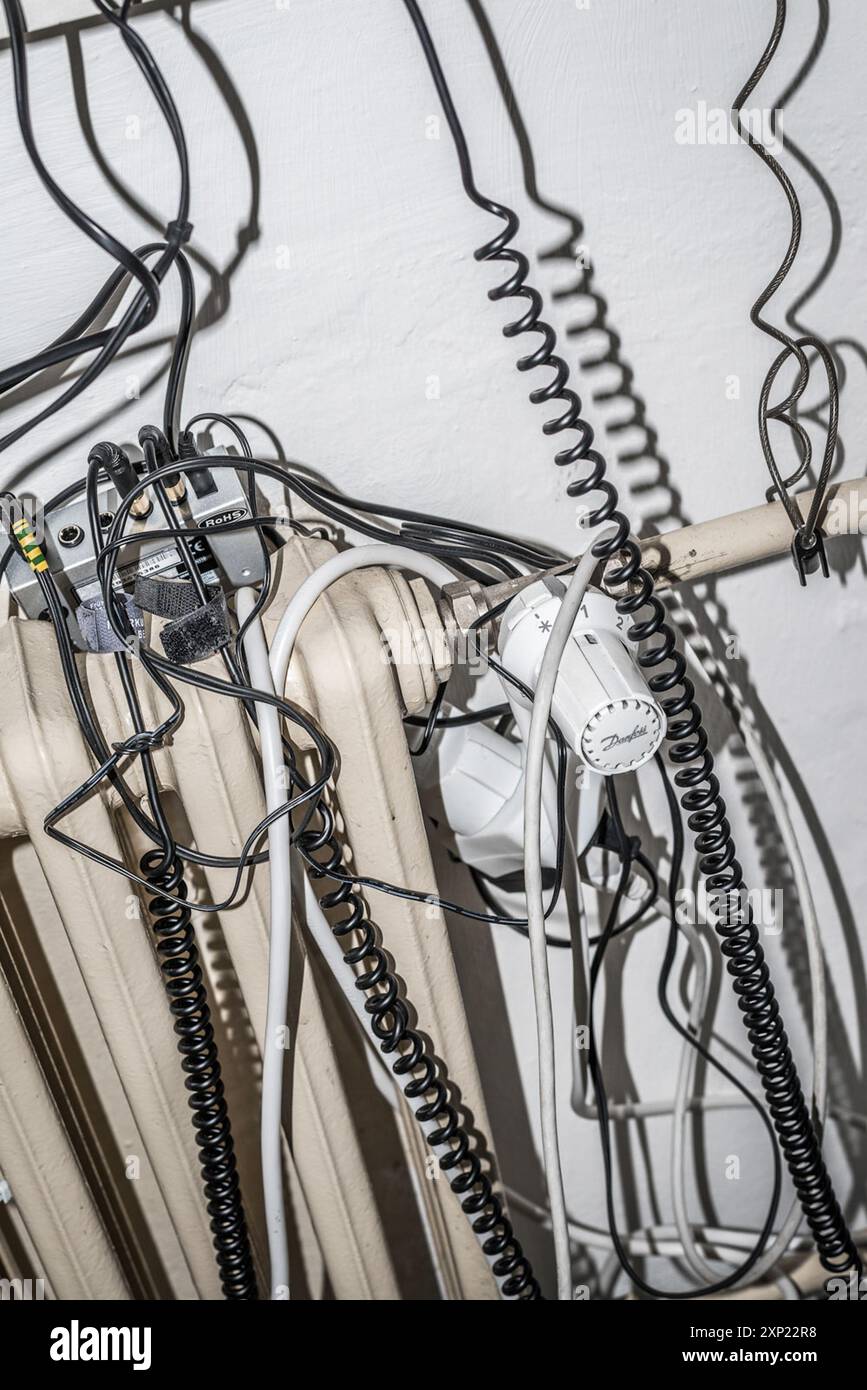 Nahaufnahme verschiedener verworrener Drähte und Kabel, die an einem Heizkörper hängen, die eine chaotische und unorganisierte Szene darstellen. Stockfoto