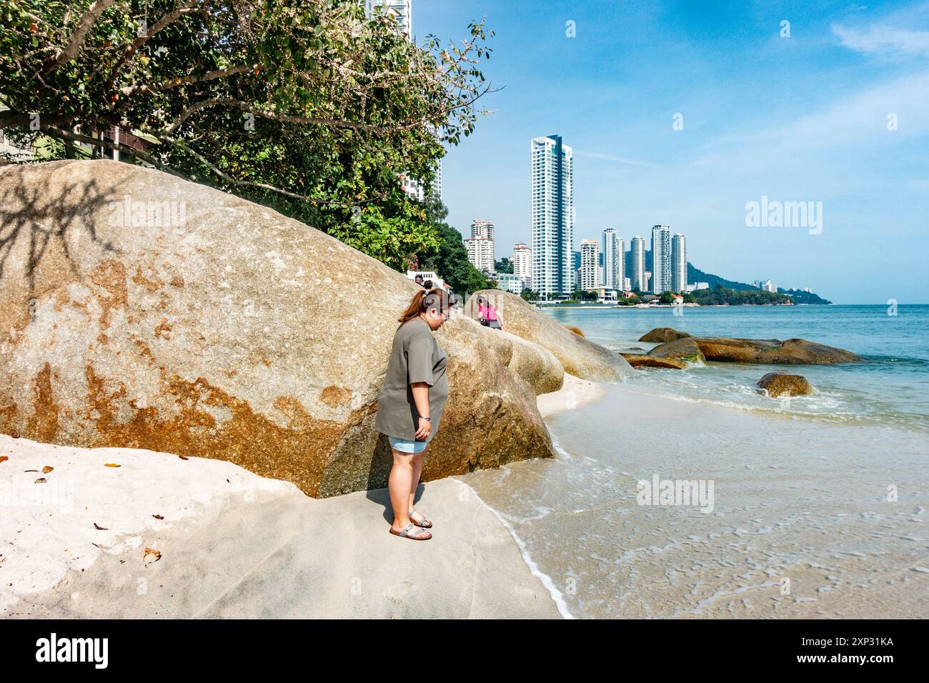 Eine Frau versucht, ihre Schuhe nicht nass zu machen am Tanjing Tokong Strand in Penang, Malaysia, mit modernen Wolkenkratzern - eine Stadt Skyscape im Hintergrund. Stockfoto