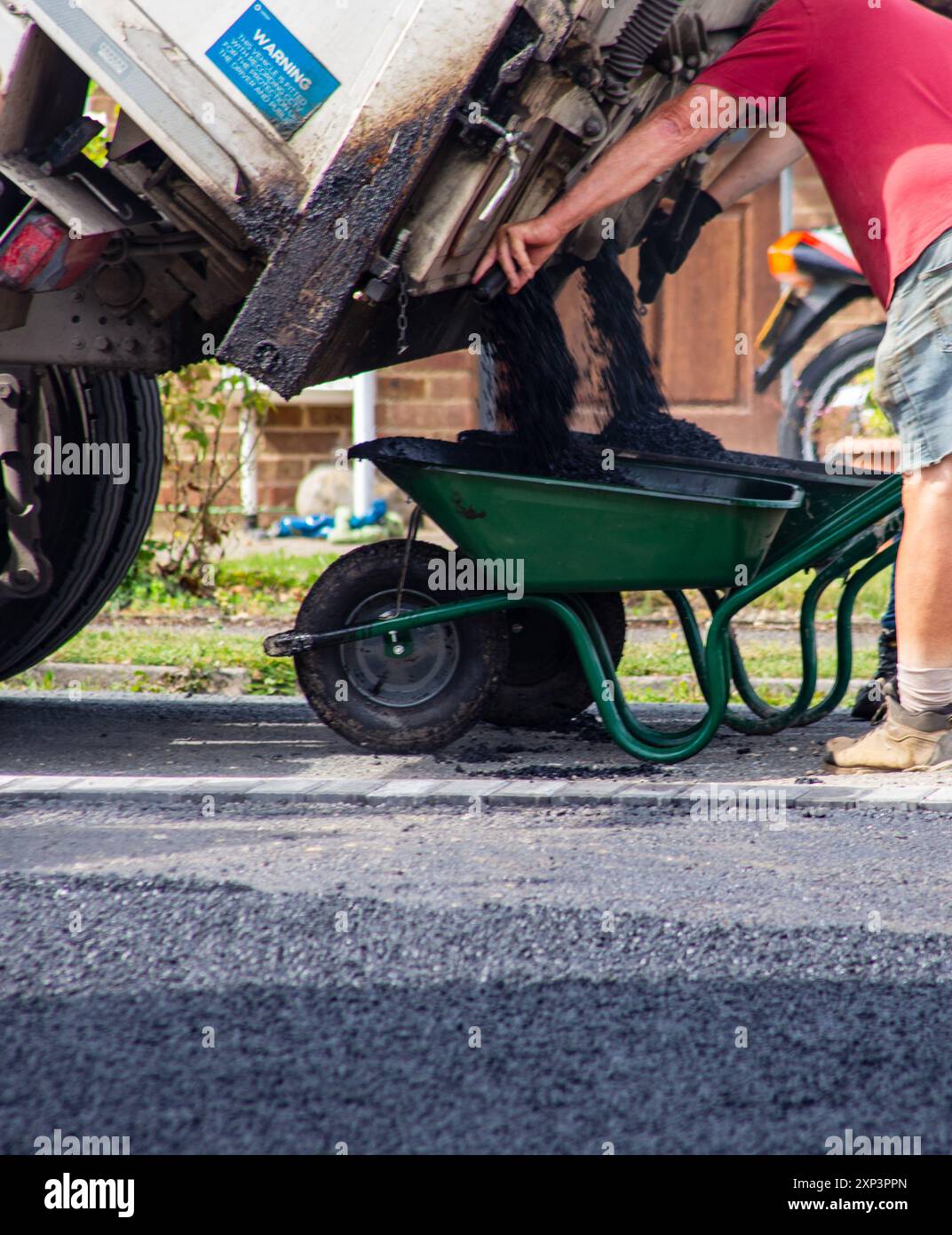 Arbeiter in rotem Hemd asphaltiert eine Straße mit frischem Teer, der aus einem Lkw in eine grüne Schubkarre gegossen wird. Stockfoto