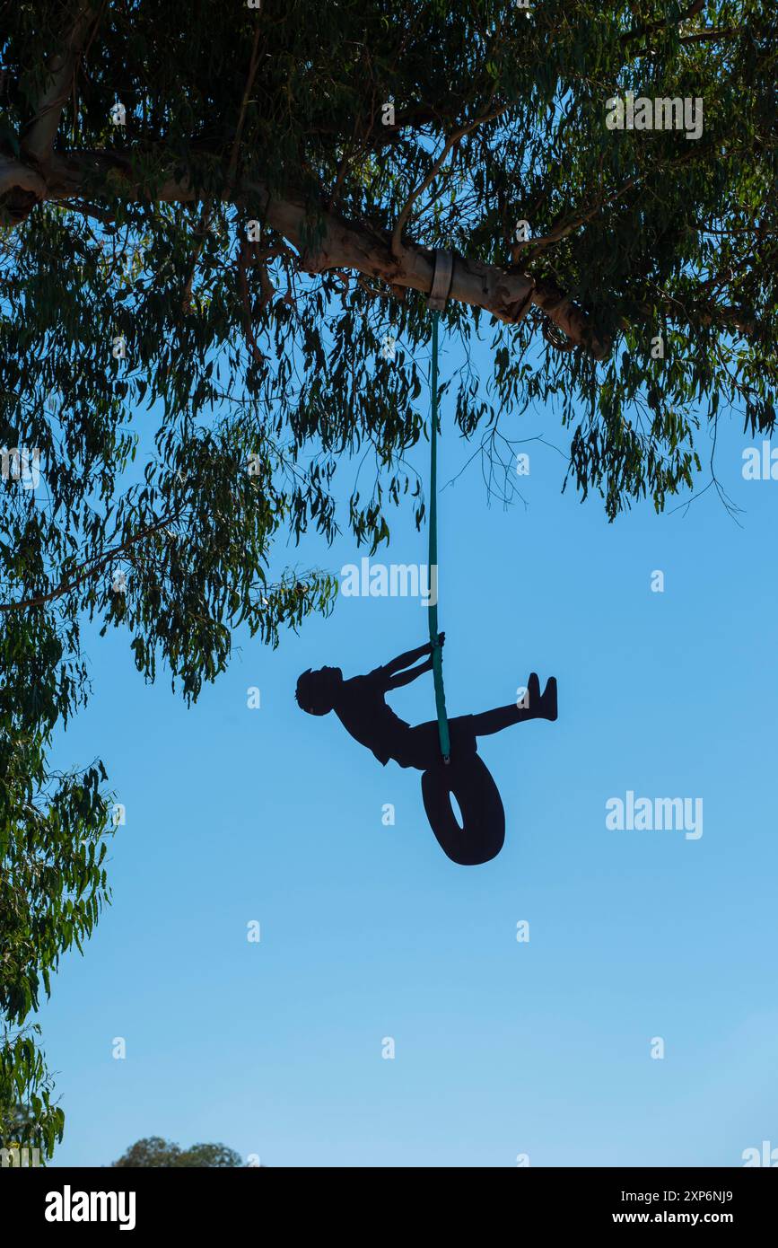 Die Skulptur „Junge auf einem Reifen“ der Künstlerin Jane Downes hängt an einem großen Baum in einem öffentlichen Park in Hastings, Neuseeland Stockfoto