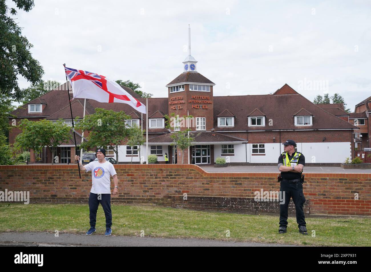 Ein einsamer Demonstrant, der von einem Polizisten während einer Demonstration vor dem Potters International Hotel in Aldershot, Hampshire beobachtet wurde, nach den Messerstechangriffen am Montag in Southport, bei denen drei kleine Kinder getötet wurden. Bilddatum: Sonntag, 4. August 2024. Stockfoto