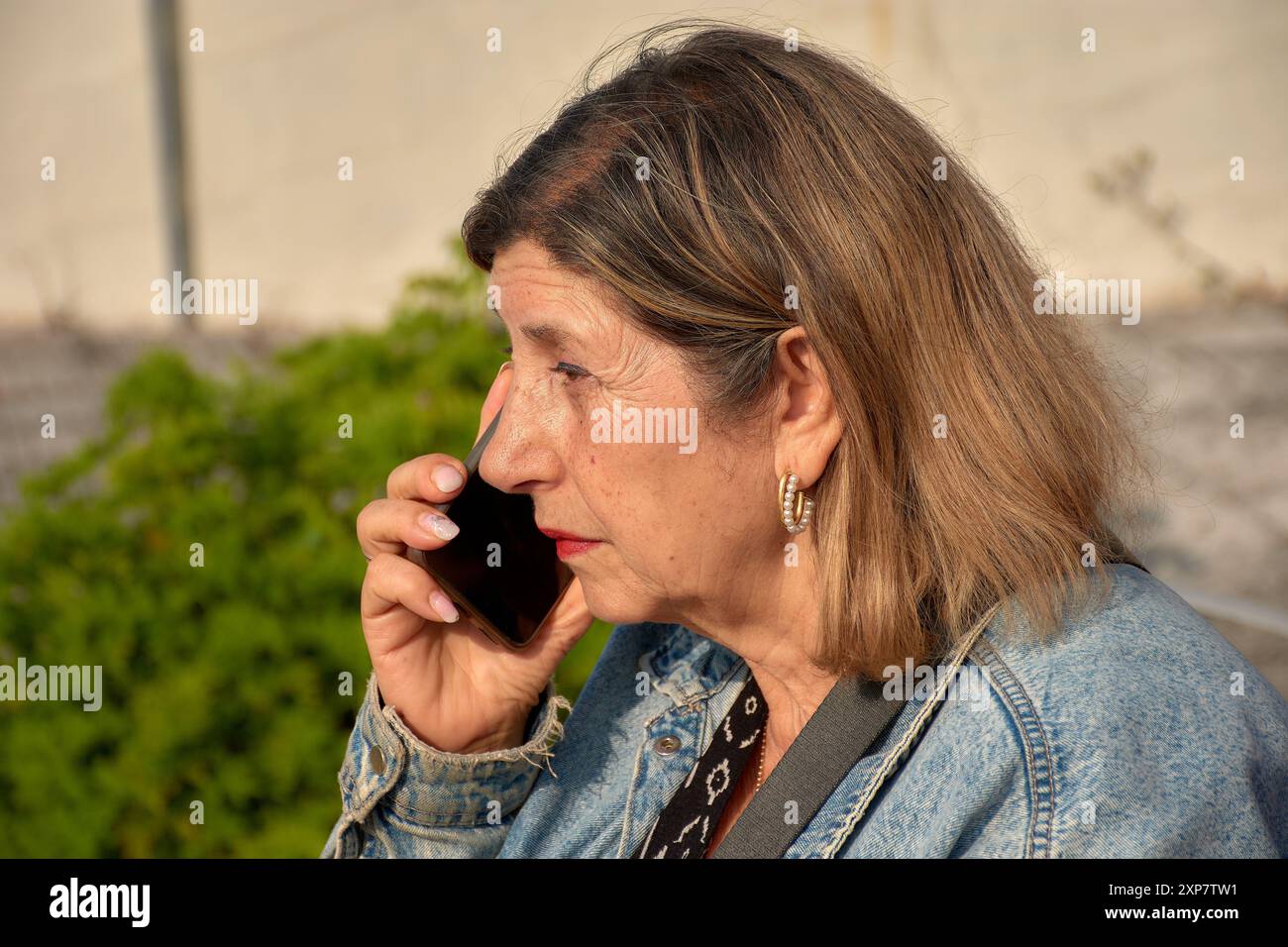 Eine ältere Frau sitzt und spricht mit einem ernsten Gesichtsausdruck auf ihrem Handy. Ihr fokussierter Blick deutet auf eine wichtige oder besorgniserregende Co hin Stockfoto