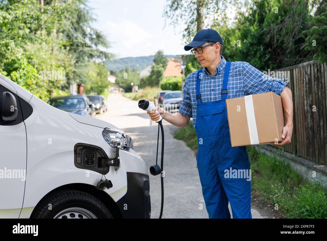 Der Liefermann in Uniform hält den Ladestecker des Elektrofahrzeugs neben dem Transporter. Stockfoto