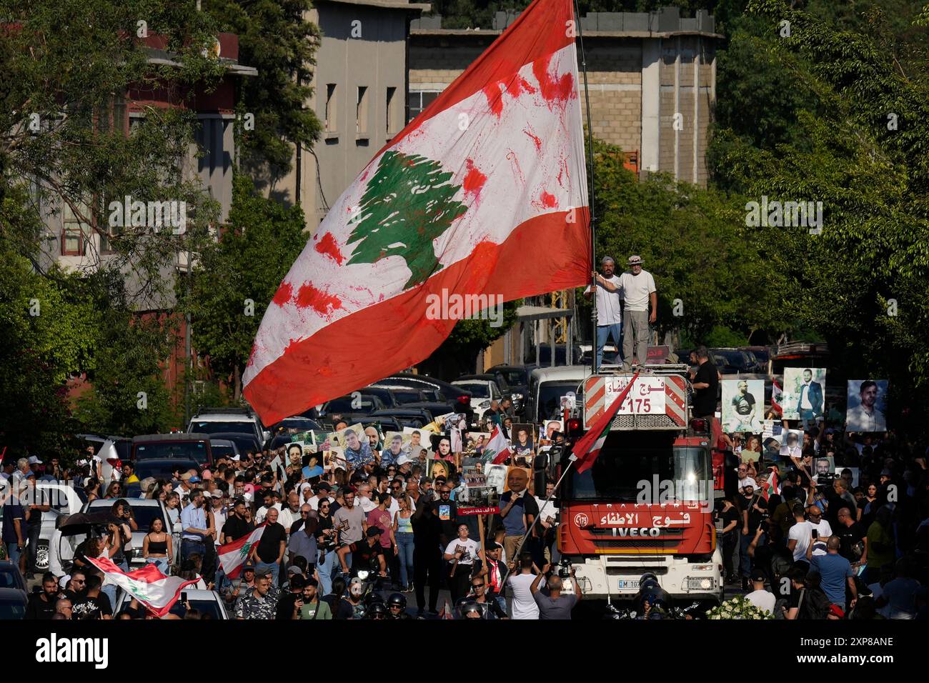 Relatives of victims who were killed in the deadly 2020 Beirut port explosion, stand on the ladder of a firetruck waving a giant Lebanese flag, as others march with portraits of loved ones during the fourth anniversary of Beirut's seaport massive blast, in Beirut, Lebanon, Sunday, Aug. 4, 2024. (AP Photo/Hussein Malla) Stockfoto