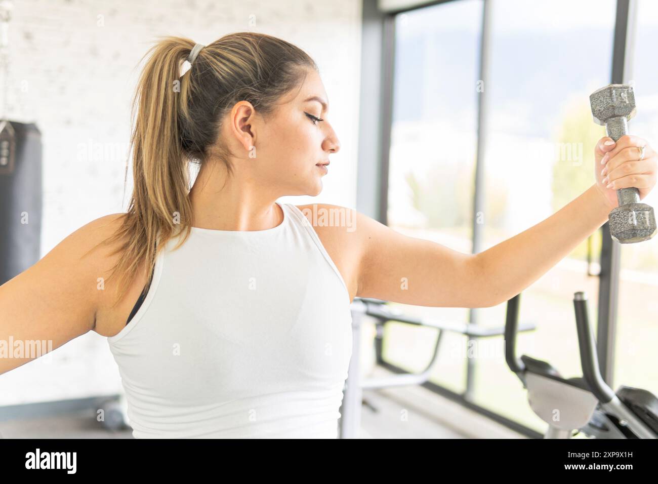 Junge lateinfrau, die auf ihren Arm schaut, während sie eine Hantel während des Trainings in einem Fitnessstudio hebt Stockfoto